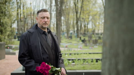 Man-In-Black-Raincoat-And-Suit-Holding-Red-Roses-Standing-In-Front-Of-A-Tombstone-In-A-Graveyard-1