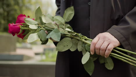 Close-Up-View-Of-Man-In-Black-Raincoat-And-Suit-Holding-Red-Roses-And-Putting-It-On-Tombstone-In-A-Graveyard
