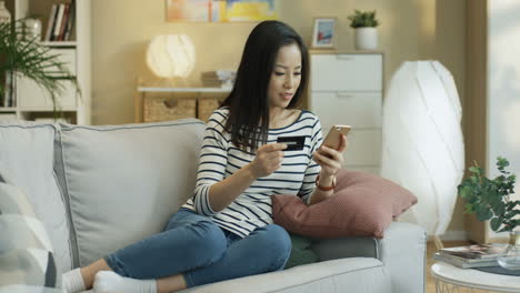 Junge-Charmante-Frau,-Die-Mit-Einem-Smartphone-Und-Einer-Kreditkarte-In-Den-Händen-Auf-Dem-Sofa-Im-Wohnzimmer-Sitzt-Und-Online-Einkauft