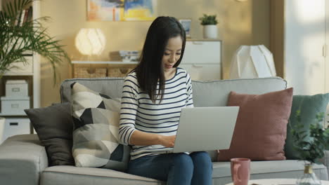 Young-Woman-Wearing-A-Striped-Blouse-And-Working-With-Laptop-Sitting-On-Sofa-In-Living-Room