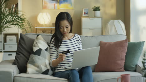 Young-Woman-Wearing-A-Striped-Blouse-With-Laptop-On-Laps-And-Shopping-Online-With-Credit-Card-Sitting-On-Sofa-In-Living-Room