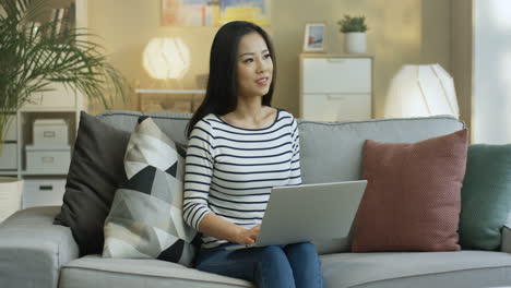 Young-Woman-In-Striped-Shirt-Working-With-Laptop-On-Laps-And-Thinking-Sitting-On-The-Sofa-In-The-Living-Room-1
