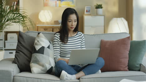 Young-Woman-In-Striped-Shirt-Working-With-Laptop-On-Laps-And-Thinking-Sitting-On-The-Sofa-In-The-Living-Room-2