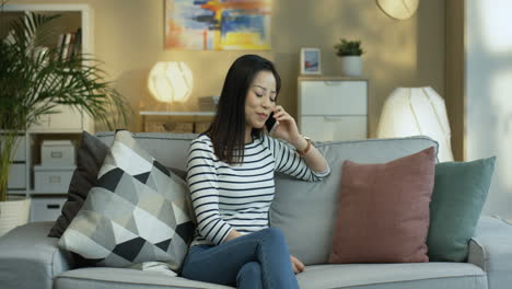 Young-Woman-In-Striped-Blouse-Sitting-On-Sofa-In-The-Living-Room-And-Talking-On-The-Phone-1