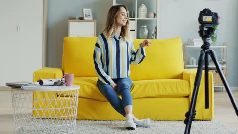 Cheerful-Female-Blogger-Talking-And-Showing-Thumbs-Up-At-Camera-While-Recording-A-Video-For-Her-Followers-Sitting-On-A-Yellow-Sofa-At-Home-1