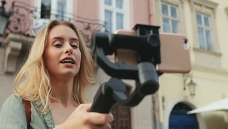 Happy-Female-Travel-Blogger-Telling-Something-While-Recording-A-Video-On-The-Smartphone-With-A-Selfie-Stick-In-The-Town