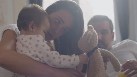 Happy-Mother-In-Bed-Holding-A-Teddy-Bear-And-Playing-With-Her-Little-Daughter-While-Dad-Watching-Them
