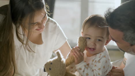 Happy-Parents-In-Bed-On-Sunday-Morning-Playing-With-Their-Cute-Baby-Girl-Who-Holding-A-Teddy-Bear
