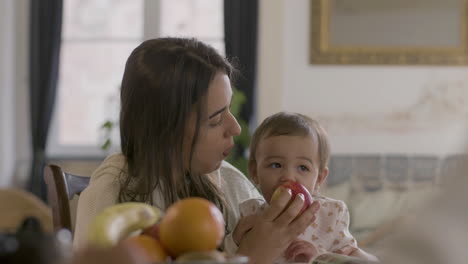 Happy-Beautiful-Woman-Sitting-At-Table-And-Holding-Her-Baby-Daughter-While-Eating-An-Apple-Together-On-The-Morning