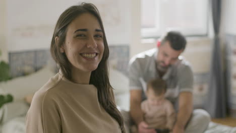 Portrait-Shot-Of-Happy-Woman-Turning-To-Camera-And-Smiling-While-In-The-Blurred-Background-Her-Husband-Sitting-On-Bed-With-Baby