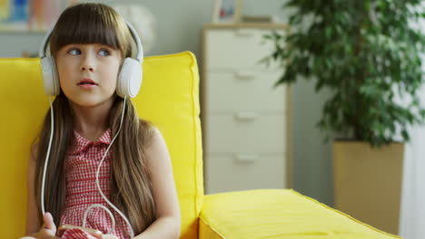 Little-Cute-Girl-Sitting-On-Yellow-Couch-Listening-To-Music-With-Headphones,-Singing-And-Dancing-Sitting-On-Sofa-In-Living-Room-At-Home