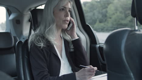 Businesswoman-Talking-On-Phone-And-Writing-Notes-In-A-Car