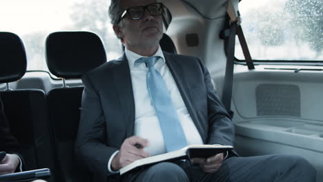 Bearded-Businessman-Writing-In-Notebook-And-Taking-Off-His-Glasses-In-A-Car