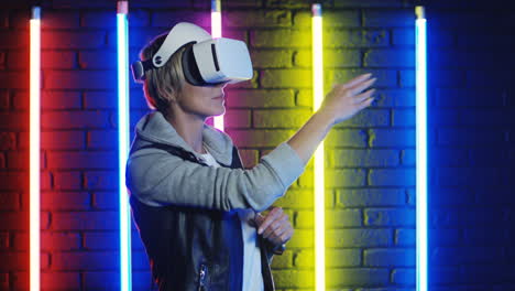 Close-Up-View-Of-Young-Blonde-Woman-In-Vr-Glasses-Moving-Her-Hands-In-The-Air-In-A-Room-With-Colorful-Neon-Lamps-On-The-Wall-1