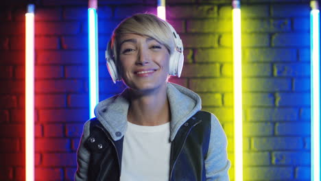 Close-Up-View-Of-Blonde-Woman-Putting-On-Big-White-Headphones-And-Listening-To-Music-In-A-Room-With-Colorful-Neon-Lamps-On-The-Wall