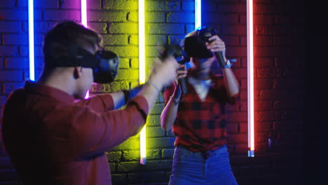 Junger-Mann-Und-Frau-In-Vr-brille-Und-Mit-Joystick-Beim-Spielen-Eines-Virtual-reality-spiels-In-Einem-Raum-Mit-Bunten-Neonlampen-An-Der-Wand-2