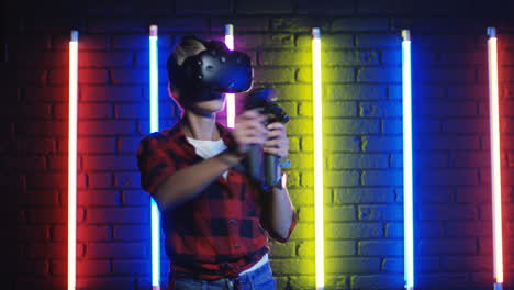 Young-Woman-In-Vr-Glasses-And-Using-Joystick-While-Playing-A-Virtual-Reality-Game-In-A-Room-With-Colorful-Neon-Lamps-On-The-Wall