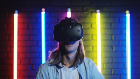Close-Up-View-Of-Young-Man-Looking-At-Camera,-Then-Put-Vr-Glasses-On-And-Starting-To-Play-A-Virtual-Game-In-A-Room-With-Colorful-Neon-Lamps-On-The-Wall