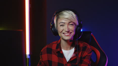 Young-Woman-With-Short-Blond-Hair-In-Big-Headphones-Sitting-In-The-Gamer-Chair-In-Front-Of-The-Computer-Screen-And-Smiling-To-The-Camera-In-A-Room-With-Colorful-Neon-Lamps-On-The-Wall