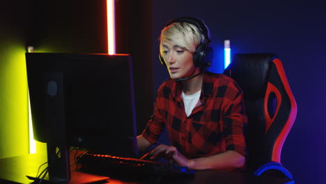 Young-Woman-With-Short-Blond-Hair-In-Big-Headphones-Sitting-In-The-Gamer-Chair-Playing-A-Game-On-The-Computer-In-A-Room-With-Colorful-Neon-Lamps-On-The-Wall