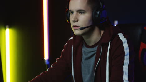 Close-Up-View-Of-The-Young-Serious-Man-With-Headphones-And-Playing-A-Game-On-The-Computer-In-A-Room-With-Colorful-Neon-Lamps-On-The-Wall