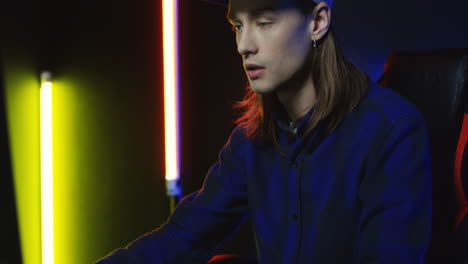 Close-Up-View-Of-Young-Serious-Man-Wearing-Cap-And-Playing-A-Game-On-The-Computer-In-A-Room-With-Colorful-Neon-Lamps-On-The-Wall