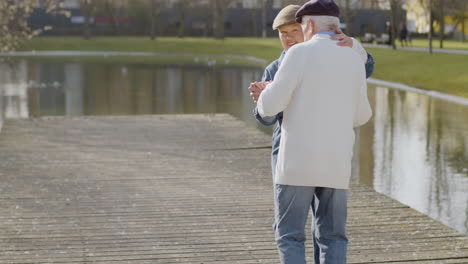 Elderly-Couple-Dancing-At-Pier-In-City-Park-On-Warm-Autumn-Day-With-Multi-Storey-Buildings-In-Background-1