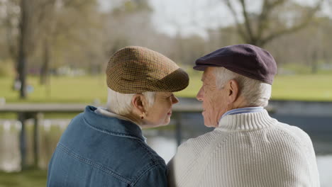 Back-View-Of-An-Elderly-Couple-Kissing-While-Sitting-On-Bench-In-Park-At-Pond-On-Nice-Autumn-Day
