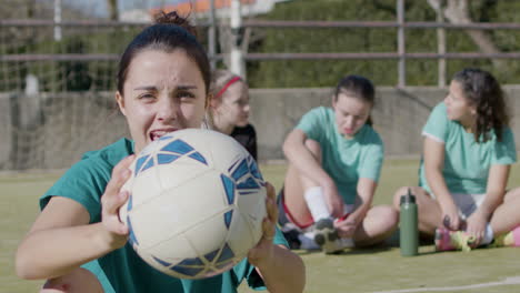 Happy-Teenaged-Girl-Holding-Football-In-Front-Of-Camera,-While-Her-Teammates-Relax-Behind