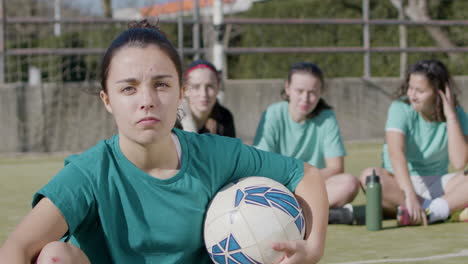 Sporty-Teenaged-Girl-Holding-Football-In-Front-Of-Camera,-While-Her-Teammates-Relax-Behind