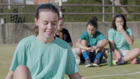 Happy-Teenaged-Girl-Holding-Football-In-Front-Of-Camera,-While-Her-Teammates-Relax-Behind-1