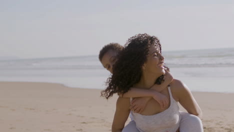 Beautiful-Young-American-Mother-Carrying-Her-Little-Son-On-Her-Back-While-Spending-Time-At-The-Beach-On-A-Sunny-Day