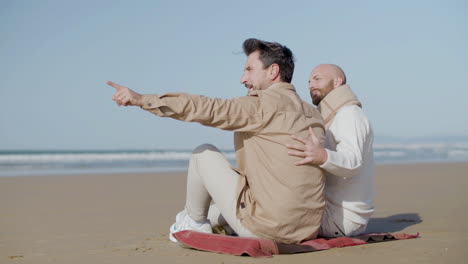 Happy-Gay-Man-Sitting-On-The-Sandy-Beach-With-His-Partner-And-Pointing-Finger-Into-Distance