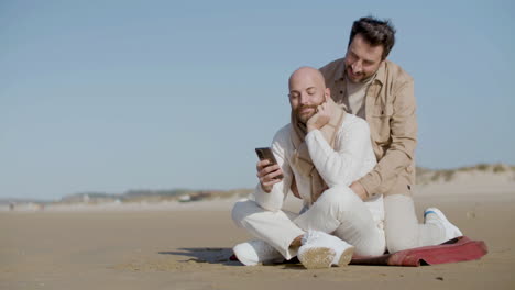 Homosexueller-Mann-Sitzt-Am-Strand-Und-Benutzt-Handy,-Während-Sein-Partner-Sich-Nähert-Und-Ihn-Von-Hinten-Umarmt