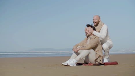 Homosexual-Man-Sitting-On-The-Beach-While-His-Partner-Approaching,-Covering-His-Eyes-And-Hugging-Him-From-Back