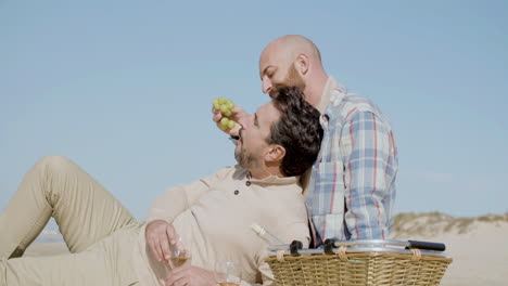 Medium-Shot-Of-A-Happy-Gay-Couple-Eating-Grape-While-Sitting-On-The-Beach