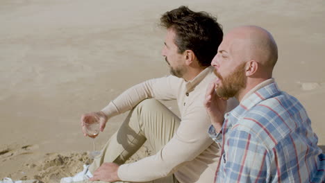 Glückliches-Schwules-Paar,-Das-Wein-Trinkt-Und-Croissants-Isst,-Während-Es-Am-Strand-Sitzt