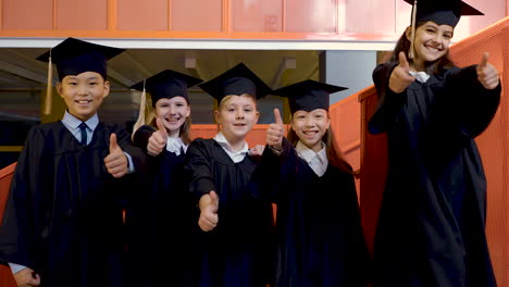 Zoom-In-Of-Happy-Kindergarten-Students-In-Cap-And-Gown-Looking-At-The-Camera-And-Doing-Thumb-Up-Sign-During-The-Preschool-Graduation-Ceremony