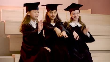 Three-Happy-Little-Girls-In-Cap-And-Gown-Playing-Together-While-Sitting-On-Stairs-At-The-Preschool-Graduation-Ceremony