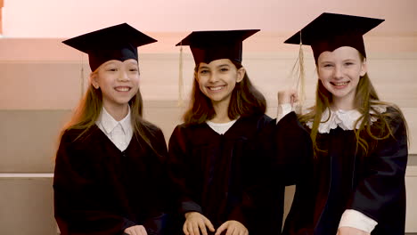 Happy-Kindergarten-Students-In-Cap-And-Gown-Smiling-And-Looking-At-The-Camera-While-Sitting-On-Stairs-During-The-Preschool-Graduation-Ceremony