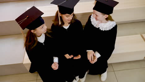 Three-Happy-Little-Girls-In-Cap-And-Gown-Sitting-On-Stairs-And-Talking-Together-At-The-Preschool-Graduation-Ceremony