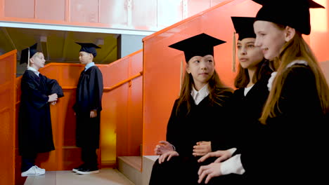 Three-Happy-Little-Girls-In-Cap-And-Gown-Sitting-On-Stairs-And-Saying-Hello-To-Their-Classmates-At-The-Preschool-Graduation-Ceremony