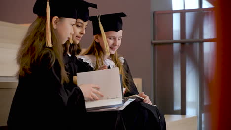 Side-View-Of-Three-Happy-Little-Girls-In-Cap-And-Gown-Talking-And-Looking-Their-Notebooks-While-Sitting-On-Stairs-At-The-Preschool-Graduation-Ceremony