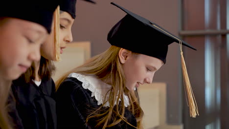 Close-Up-Of-Three-Happy-Little-Girls-In-Cap-And-Gown-Talking-Together-While-Sitting-On-Stairs-At-The-Preschool-Graduation-Ceremony