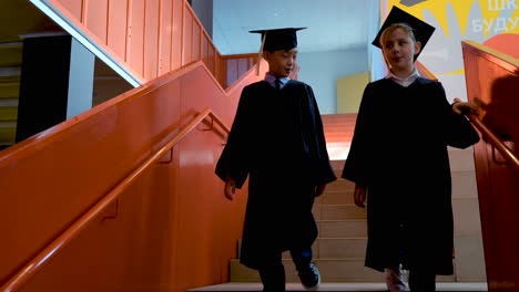 Two--Kindergarten-Students-In-Cap-And-Gown-Talking-Together-While-Going-Down-The-Stairs-At-The-Preschool-Graduation-Ceremony