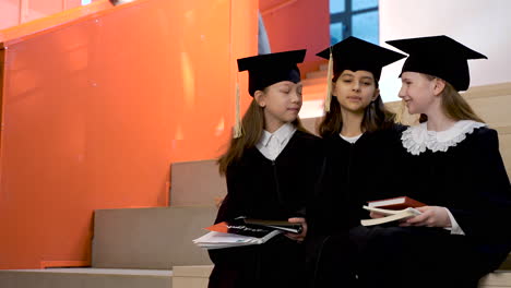Three-Happy-Little-Girls-In-Cap-And-Gown-Talking-And-Holding-Their-Notebooks-While-Sitting-On-Stairs-At-The-Preschool-Graduation-Ceremony
