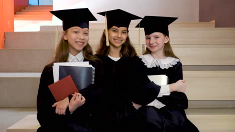 Portrait-Of-Three-Happy-Little-Girls-In-Cap-And-Gown-Holding-Notebooks-And-Looking-At-The-Camera-While-Sitting-On-Stairs-At-The-Preschool-Graduation-Ceremony