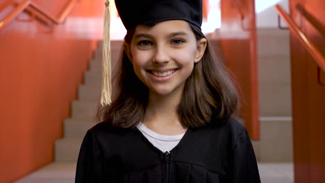 Portrait-Of-A-Happy-Preschool-Female-Student-In-Cap-And-Gown-Going-Down-The-Stairs,-Holding-Graduation-Diploma-And-Looking-At-The-Camera