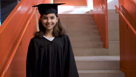 Portrait-Of-A-Happy-Preschool-Female-Student-In-Cap-And-Gown-Going-Down-The-Stairs,-Holding-Graduation-Diploma-And-Looking-At-The-Camera-1