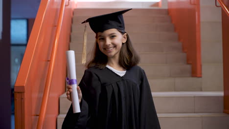 Portrait-Of-A-Happy-Preschool-Female-Student-In-Cap-And-Gown-Running-Down-The-Stairs,-Holding-Graduation-Diploma-And-Looking-At-The-Camera